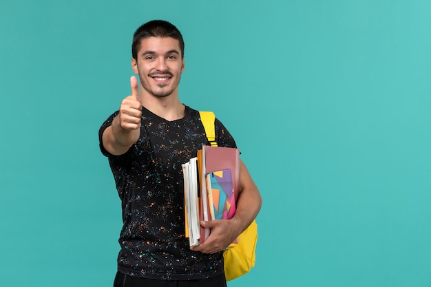 Front view of male student in dark t-shirt wearing yellow backpack holding copybook and files smiling on blue wall