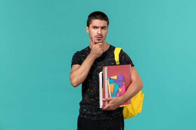 Студент в темной футболке с желтым рюкзаком, держащий тетрадь и файлы, думающий на синей стене, вид спереди
