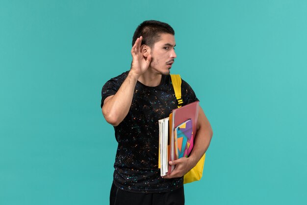 青い壁で聞こうとしているコピーブックとファイルを保持している黄色のバックパックを身に着けている暗いTシャツの男子学生の正面図