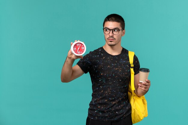 Вид спереди студента в темной футболке с желтым рюкзаком, держащего кофе и часы на синей стене