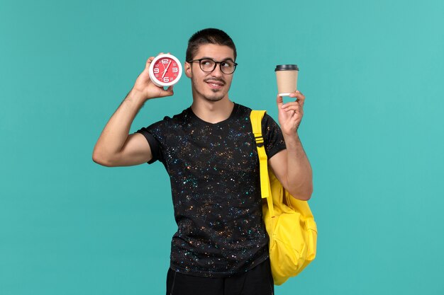 Front view of male student in dark t-shirt yellow backpack holding coffee and clocks on the blue wall