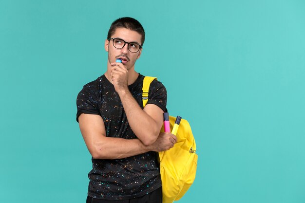 青い壁に考えている色のフェルトペンを保持している暗いTシャツ黄色のバックパックで男子学生の正面図