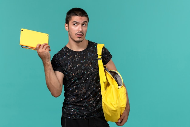 Free photo front view of male student in dark t-shirt yellow backpack holding copybook on light blue wall