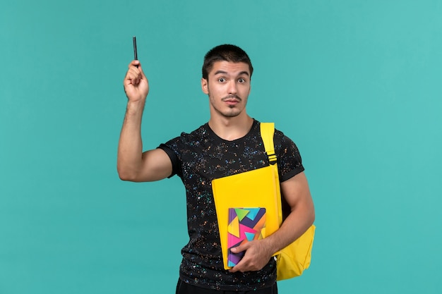 Вид спереди студента в темной футболке с желтым рюкзаком, держащего ручку для тетради и файлы на голубой стене