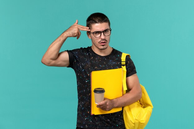 Front view of male student in dark t-shirt yellow backpack holding different files and coffee on blue wall