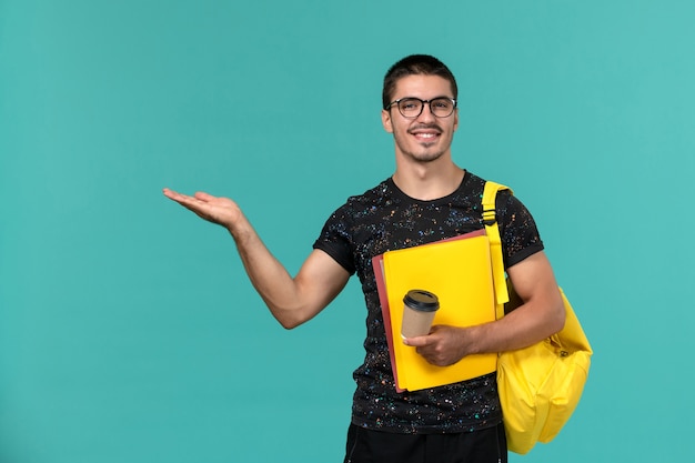 Front view of male student in dark t-shirt yellow backpack holding different files and coffee smiling on blue wall