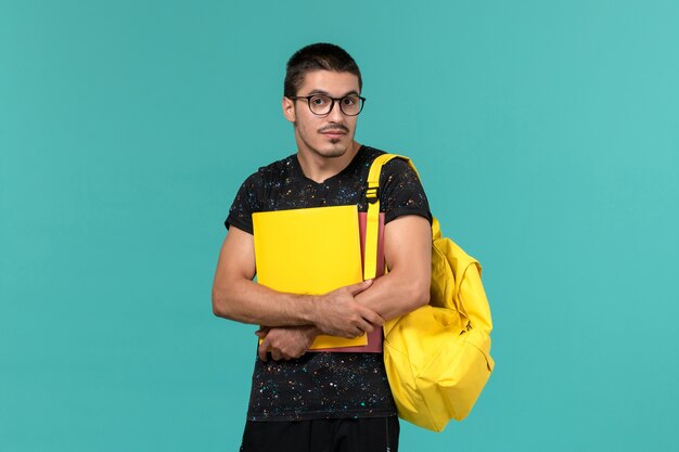 Студент в темной футболке с желтым рюкзаком, держащий разные файлы на голубой стене, вид спереди