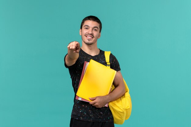 Студент в темной футболке с желтым рюкзаком, держащий разные файлы на голубой стене, вид спереди