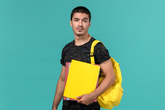 밝은 파란색 벽에 다른 파일을 들고 어두운 티셔츠 노란색 배낭에 남성 학생의 전면보기
