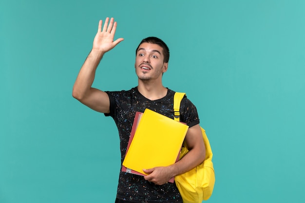 Вид спереди студента в темной футболке с желтым рюкзаком с разными файлами, размахивающего на голубой стене