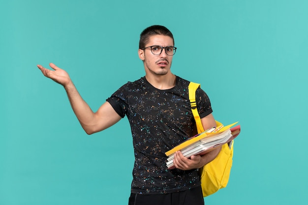 水色の壁にファイルと本を保持している暗いTシャツ黄色のバックパックで男子学生の正面図