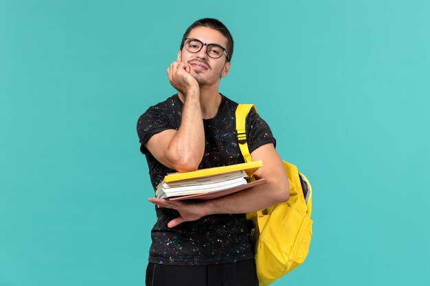 水色の壁にファイルと本を保持している暗いTシャツ黄色のバックパックで男子学生の正面図
