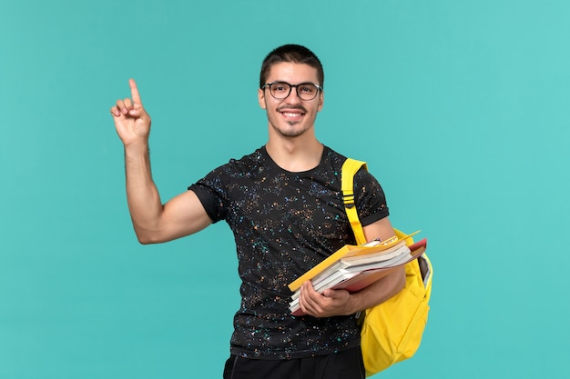 Front view of male student in dark t-shirt yellow backpack holding files and books on light blue wall