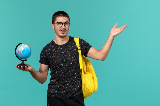 Студент в темной футболке с желтым рюкзаком, держащий глобус на синей стене, вид спереди