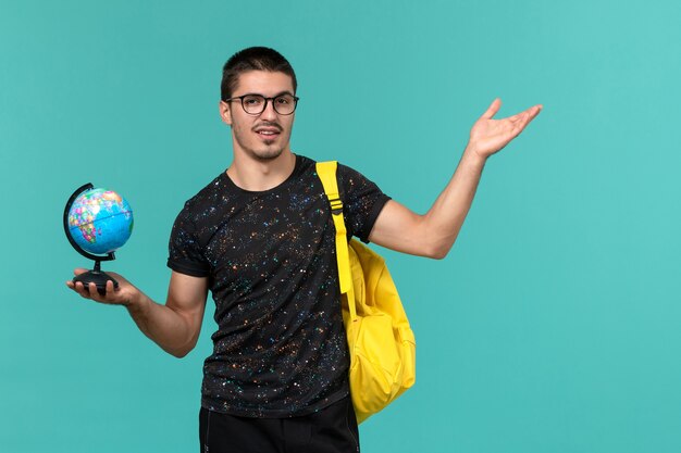 青い壁に小さな地球を保持している暗いTシャツ黄色のバックパックで男子学生の正面図