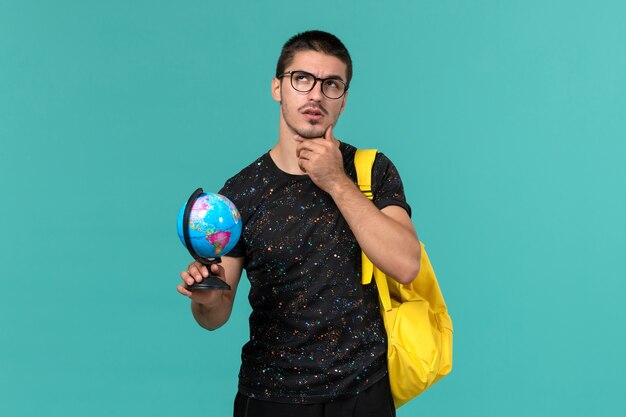 Front view of male student in dark t-shirt yellow backpack holding little globe and thinking on blue wall