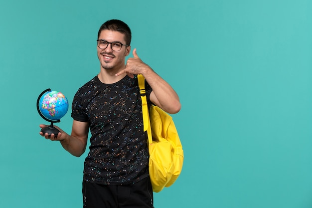 Вид спереди студента в темной футболке с желтым рюкзаком, держащего маленький глобус с улыбкой на синей стене
