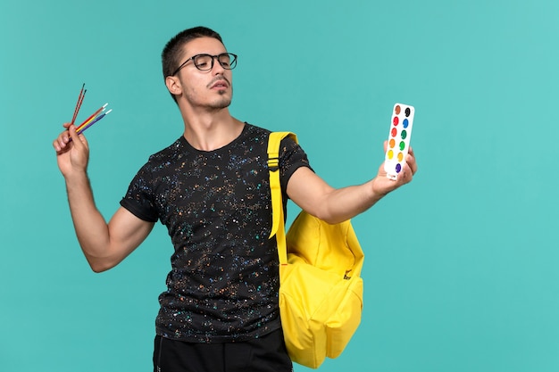 Front view of male student in dark t-shirt yellow backpack holding paints on light blue wall