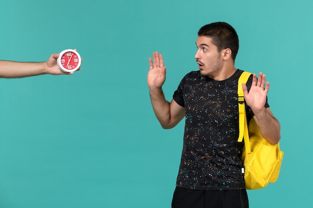 Front view of male student in dark t-shirt yellow backpack looking at clocks on light blue wall