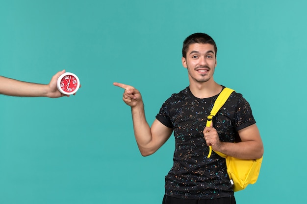 Front view of male student in dark t-shirt yellow backpack smiling on light blue wall