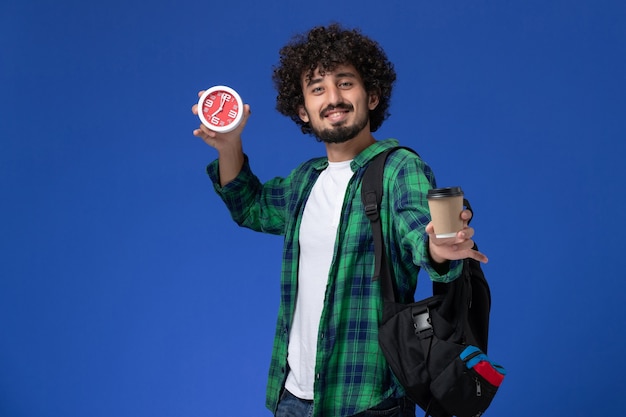 Front view of male student in green checkered shirt wearing black backpack and holding clocks and coffee on blue wall