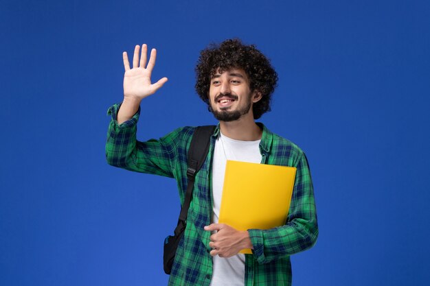 Front view of male student in green checkered shirt wearing black backpack and holding files on blue wall