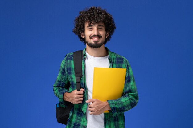 Front view of male student in green checkered shirt wearing black backpack and holding files on blue wall