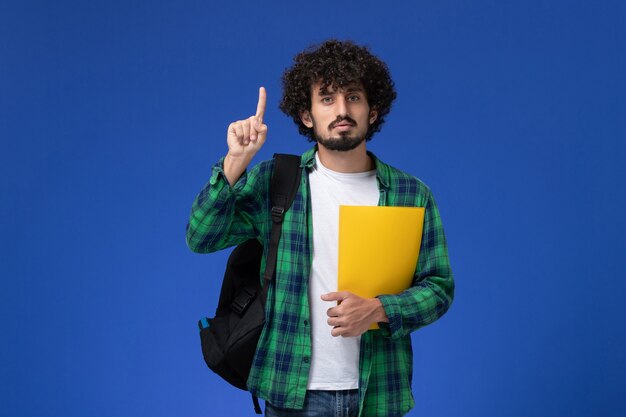 Front view of male student in green checkered shirt wearing black backpack and holding files on the blue wall