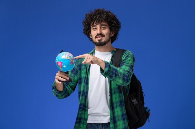 Front view of male student in green checkered shirt wearing black backpack and holding little globe on blue wall