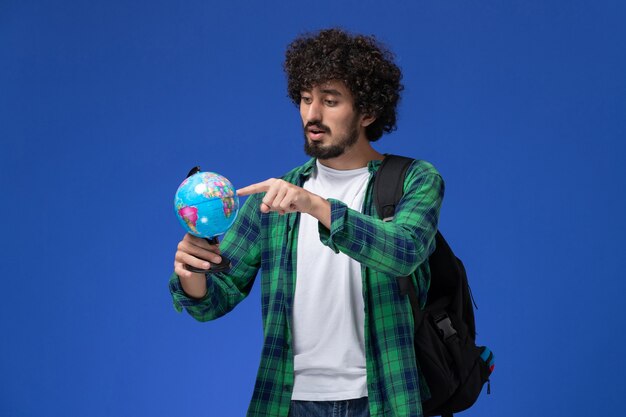 Front view of male student in green checkered shirt wearing black backpack and holding little globe on blue wall