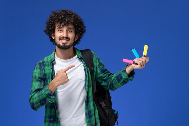 Front view of male student in green checkered shirt with black backpack holding colored felt pen on blue wall