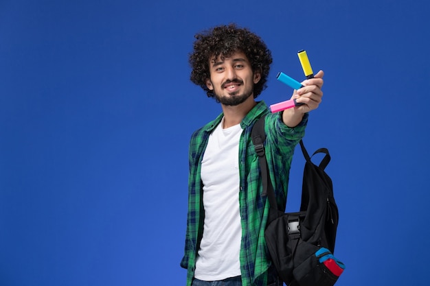 Front view of male student in green checkered shirt with black backpack holding colored felt pens on blue wall