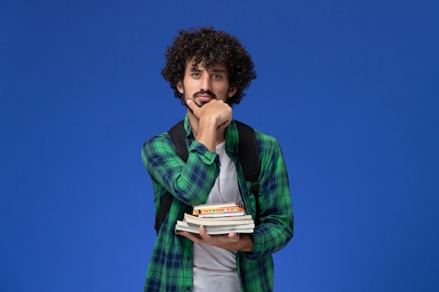 파란색 벽에 카피 북 및 파일을 들고 검은 배낭과 녹색 체크 무늬 셔츠에 남성 학생의 전면보기