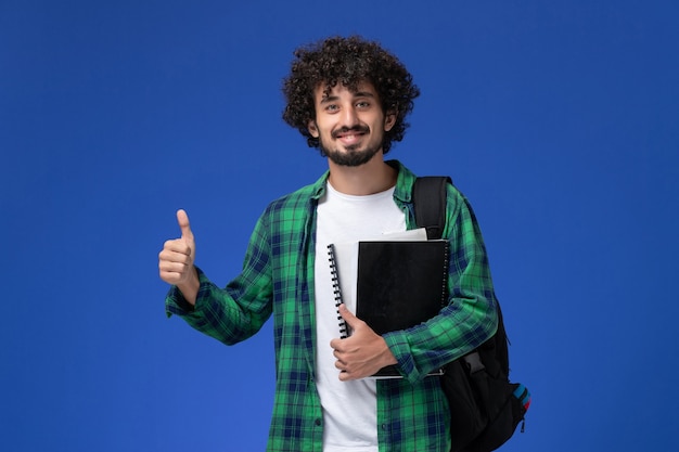 Vista frontale dello studente maschio in camicia a scacchi verde con zaino nero che tiene i quaderni sorridente sulla parete blu