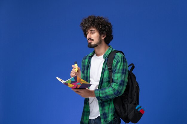 Front view of male student in green checkered shirt with black backpack holding felt pen and copybook on blue wall