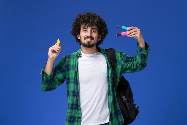 Front view of male student in green checkered shirt with black backpack holding felt pens on blue wall