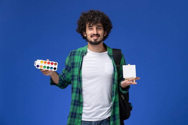 Front view of male student in green checkered shirt with black backpack holding paints and easel on blue wall