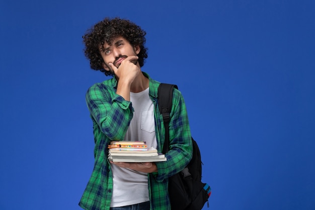 Foto gratuita vista frontale di uno studente maschio in camicia a scacchi verde con zaino nero che pensa tenendo i quaderni e file sulla parete blu