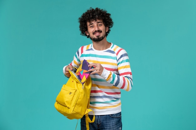 Front view of male student holding yellow backpack and copybook on blue wall