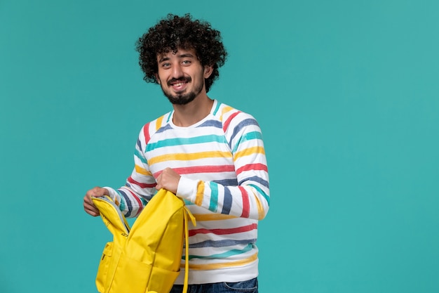 Vista frontale dello studente maschio che tiene zaino giallo e quaderno sorridente sulla parete blu