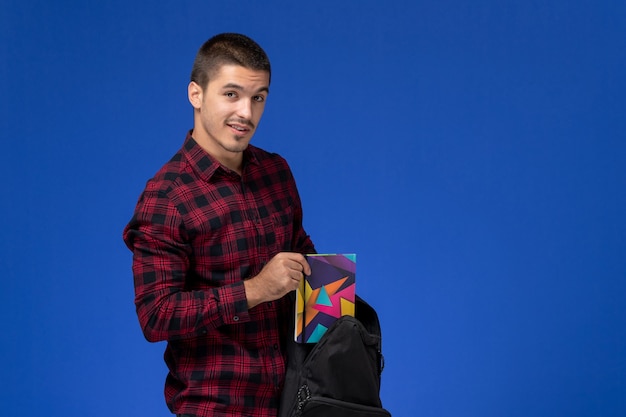 Front view of male student in red checkered shirt holding his backpack and copybook on the blue wall