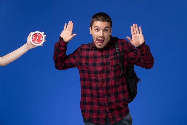 Front view of male student in red checkered shirt with backpack on the blue wall