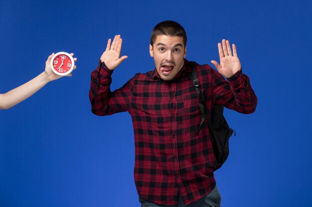 Front view of male student in red checkered shirt with backpack on the blue wall