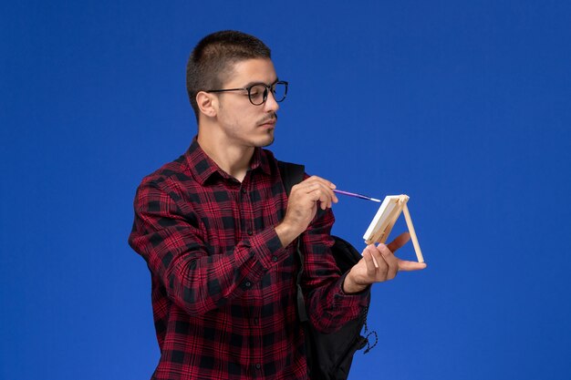 Front view of male student in red checkered shirt with backpack drawing on easel on blue wall