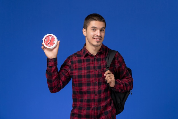 Front view of male student in red checkered shirt with backpack holding clocks on blue wall