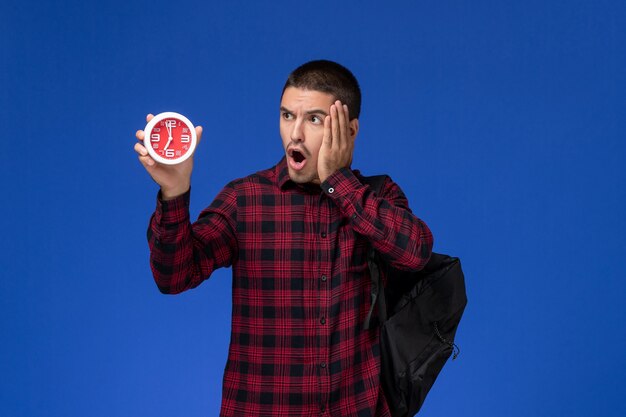 Front view of male student in red checkered shirt with backpack holding clocks on blue wall