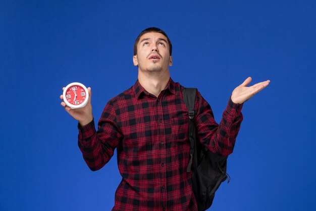 Вид спереди студента в красной клетчатой рубашке с рюкзаком, держащим часы на синей стене