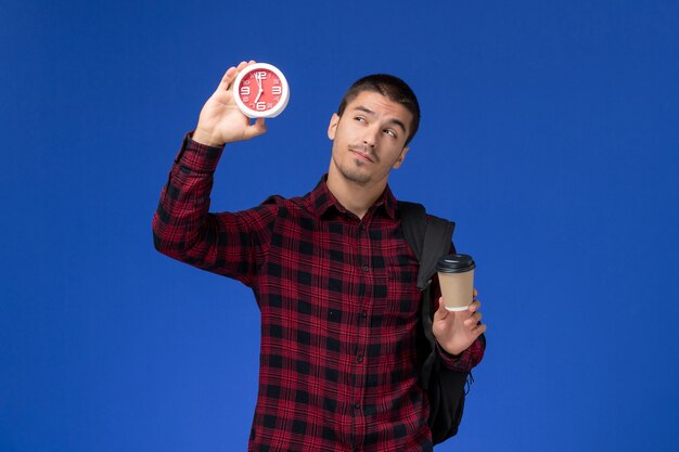 Front view of male student in red checkered shirt with backpack holding clocks and coffee on blue wall