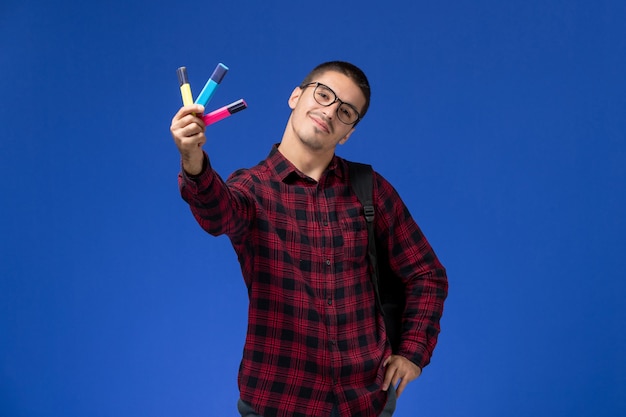 Front view of male student in red checkered shirt with backpack holding colorful felt pens on light-blue wall