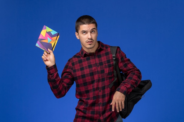 Front view of male student in red checkered shirt with backpack holding copybook on blue wall